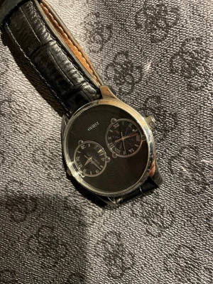 Svart läderklocka från Guess - Stilren klocka från Guess med svart urtavla och dubbla tidzoner. Boetten är i rostfritt stål och armbandet är gjort av svart läder med struktur. Urtavlan har både vanliga siffror och romerska siffror för en unik look. Perfekt accessoar för dig som gillar klassisk stil.