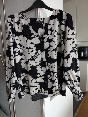 Svartvit blommig blus från Stockh LM - Snygg blus med svart botten och vita blommönster från Stockh LM. Blusen har v-ringning, knappar framtill och långa ärmar med knäppning vid mudden. Materialet är lätt och luftigt, perfekt för att skapa en cool och trendig look.