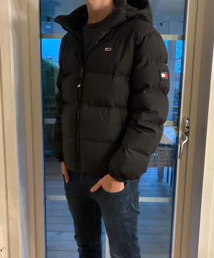 Svart pufferjacka från Tommy Hilfiger - Säljer denna väldigt sköna svart pufferjackan från Tommy Hilfiger med huva och dragkedja framtill. Jackan har logotyp på bröstet och ärmen, samt broderad text på insidan. Modellen på bilden är: 186 och väger 72. HELT NY! Perfekt för kalla dagar. Pris kan diskuteras!
