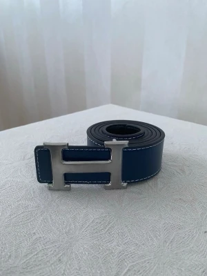 Blått skärp med H-spänne  - Snyggt blått skärp med ikoniskt H-format metallspänne. Skärpet har vita kontrastsömmar längs kanterna och är tillverkat i slätt skinn. Perfekt accessoar för att lyfta din outfit.