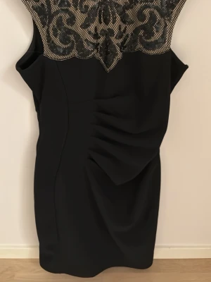 Svart kort klänning med spetsdetalj i strl 42 - Elegant svart kort klänning med vackert mönstrad spets upptill i beige och svart. Klänningen har drapering framtill och är ärmlös med djup v-ringning i ryggen. Perfekt för dig som vill sticka ut med stilrena detaljer.