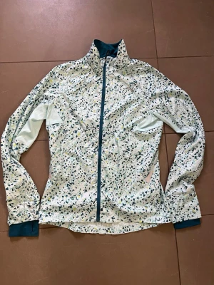 Mönstrad vindjacka från Puma - Snygg vindjacka från Puma med vitt tyg och abstrakt mönster i blått, grönt och gult. Jackan har hel dragkedja framtill, hög krage och muddar i mörkblått. Tillverkad i lätt och funktionellt material som passar perfekt för träning utomhus.