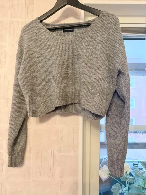 Croppad stickad tröja - Säljer en jättefin grå stickad tröja i croppad modell. Tröjan har en snygg ribbstickad textur, vid V-ringning och är något längre baktill än framtill. Har mycket stretch och passformen är avslappnad. Katt finns i hemmet.   Material: 62% polyester, 34% akryl och 4% elastan Nypris: 278kr. Mitt pris: 70kr. Spara 208kr!