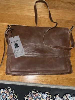 Brun axelväska i äkta läder - Snygg brun axelväska i äkta handgjort original läder med justerbar axelrem. Väskan har ett stort huvudfack, innerfack med dragkedja och magnetlås. Perfekt för dig som gillar klassisk och stilren design med praktiska fack för dina grejer.