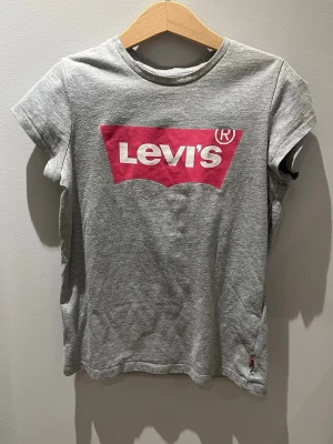 Grå Levi's t-shirt med logga - Snygg grå t-shirt från Levi's med den klassiska röda loggan tryckt på bröstet. T-shirten har korta ärmar och rund halsringning. Tillverkad i mjuk bomull för en skön känsla. Perfekt till jeans eller shorts för en avslappnad look.
