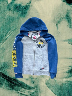 Superdry blå och grå hoodie - Säljer denna väldigt feta och unika hoodien från Superdry! Ascoolt plagg med olika färger från armarna och kroppen. Väldigt snygg sommarhoodie som man kan ha under sena sommarkvällar. Hör av dig!