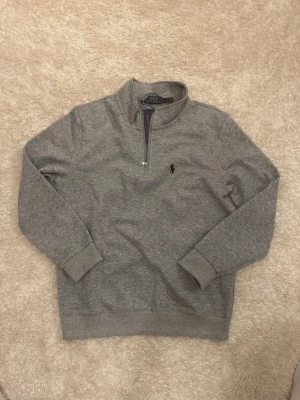 Grå halvzip tröja från Polo Ralph Lauren - Klassisk gråmelerad sweatshirt från Polo Ralph Lauren med halv dragkedja och broderad logga på bröstet. Tröjan har långa ärmar, ribbade muddar och är tillverkad i mjuk bomullsblandning. Perfekt för en avslappnad och stilren look. Står storlek L men skulle nog säga storlek S/Xs passar bättre 