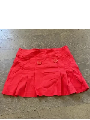 Röd kortkjol med knappar - Säljer en snygg röd lågmidjad y2k kortkjol med två dekorativa knappar framtill och plisserad nederkant. Kjolen är tillverkad i bomull och har en ungdomlig och fräsch look som passar perfekt till varma dagar.