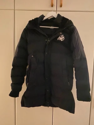 Svart dunjacka Kings Will Dream XS - Snygg svart dunjacka från Kings Will Dream i storlek XS. Jackan har huva, dragkedja och tryckknappar framtill samt två stora fickor. Märkets logga i vitt på bröstet ger en streetwear-vibe. Perfekt för kalla vinterdagar.