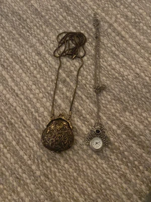 Två vintageinspirerade halsband - Säljer två halsband med unika hängen. Det ena har ett bronsfärgat, rundat hänge med snirkliga mönster och bladdekor. Det andra har ett silverfärgat hänge med klocka och svart sten, dekorerat med detaljerade mönster. Båda har långa kedjor och retrovibbar.