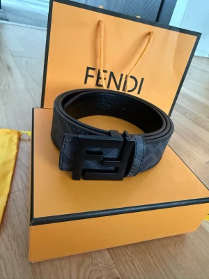 Svart bälte från Fendi med logga - Snyggt svart bälte från Fendi med klassisk Fendi-logga på det mattsvarta spännet. Bältet har diskret mönster i mörka toner och är gjort i exklusivt skinn. Perfekt accessoar för att lyfta din outfit.
