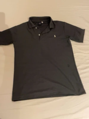 Mörkgrå pikétröja från Ralph Lauren - Klassisk mörkgrå pikétröja från Ralph Lauren med korta ärmar, krage och knappar framtill. Liten broderad logga på bröstet. Tillverkad i mjuk bomull för en bekväm känsla och stilren look.