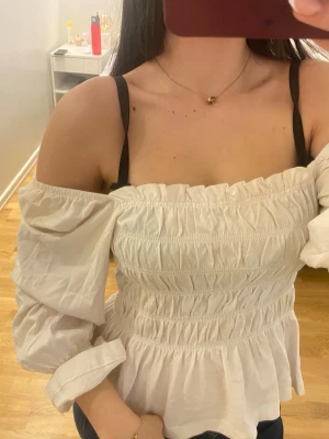 Vit off shoulder topp med puffärm - Trendig vit off shoulder topp med smockad överdel och vida puffärmar. Toppen har en rynkad, elastisk passform och volangkant nedtill. Perfekt för dig som gillar romantisk och feminin stil. Materialet ser ut att vara bomull eller bomullsblandning.