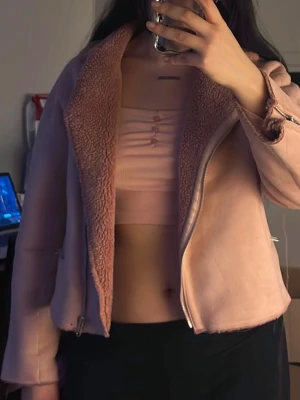 Ljusrosa teddyfodrad jacka - Säljer en ljusrosa jacka med mjukt teddyfoder och dragkedja framtill. Jackan har en bred krage, detaljerade muddar med spännen och en croppad passform. Perfekt för dig som vill ha en mysig och trendig look i höst och vinter.