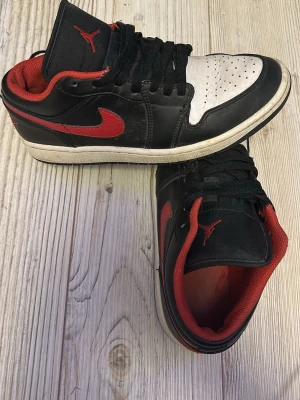 Air Jordan 1 Low svart/röd/vit - Säljer ett par Air Jordan 1 Low sneakers från Nike. Skorna är svarta med röda detaljer och vit tå, har klassisk röd swoosh och Jumpman-logga på tungan. Yttermaterialet är läder och insidan är fodrad med rött tyg. Perfekt för dig som gillar streetstyle och ikoniska sneakers.