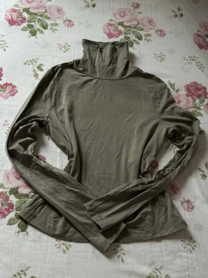 Olivgrön polotopp med långa ärmar - Säljer en olivgrön polotopp med lång ärm och hög krage. Toppen har en enkel, stilren design och är tillverkad i mjukt material som känns skönt mot huden. Perfekt att styla lager på lager eller bära som den är.