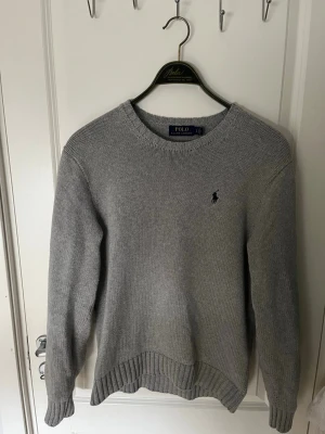Grå stickad tröja Polo Ralph Lauren - Klassisk grå stickad tröja från Polo Ralph Lauren med rund halsringning och diskret broderad logga på bröstet. Tröjan har ribbstickade muddar vid ärmslut och nederkant. Perfekt för lager på lager och enkel att matcha.
