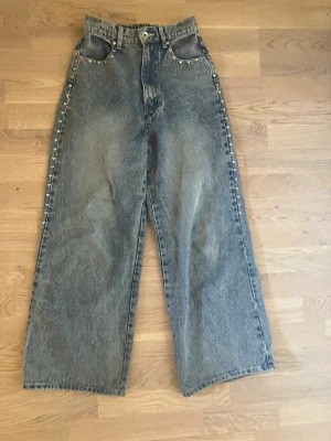 Vida blå jeans med hjärtan - Säljer ett par coola vida blå jeans med hög midja och dekorativa nitar längs fickor och sidor. Jeansen har klassisk femficksmodell och är gjorda i kraftigt denimtyg. Perfekta för dig som gillar en avslappnad och trendig stil.