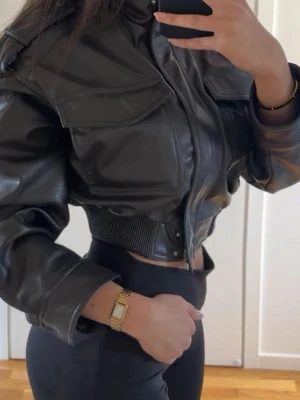 Kort brun skinnjacka  - Snygg brun skinnjacka med hög krage och dragkedja framtill. Jackan har två stora bröstfickor med lock, ribbad midja och axeldetaljer. 