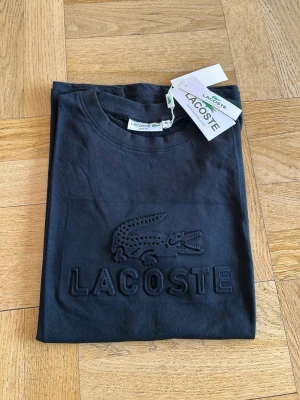 Svart Lacoste t-shirt med logga - Snygg svart t-shirt från Lacoste med stor broderad krokodil och Lacoste-text på bröstet. Klassisk rund hals och korta ärmar. Tillverkad i mjuk bomull som känns skön mot huden. Perfekt för dig som gillar stilrena och tidlösa plagg.
