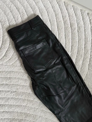 Svarta fuskskinnbyxor från Zara - Säljer ett par svarta byxor i fuskskinn från Zara. Byxorna har raka ben, hög midja och klassiska fickor både fram och bak. Finns en slits längst ned. Referens: jag är 180cm (byxorna är precis ovanför golvet)