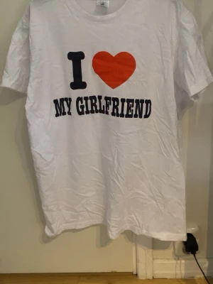 Vit t-shirt med tryck 'I ❤️ MY GIRLFRIEND' - Vit t-shirt med stort tryck på bröstet där det står 'I ❤️ MY GIRLFRIEND' i svart och rött. Klassisk passform och rund halsringning. Perfekt för att visa lite extra kärlek till din flickvän.