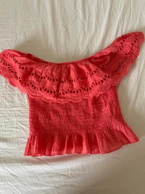 Korall offshoulder topp med volang - Trendig rosa offshoulder topp med bred volangkant och broderade detaljer. Smockad kropp och volang längst ner ger en söt och feminin look. Perfekt för varma dagar och sommarens alla tillfällen.