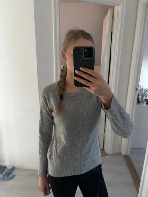 Grå kabelstickad tröja - Säljer en grå kabelstickad tröja med rund hals och långa ärmar. Tröjan har ett klassiskt mönster och ribbade muddar vid ärmslut och nederkant. 