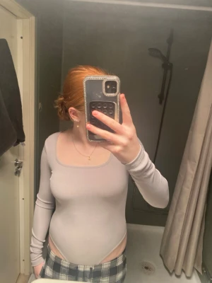 Ljusgrå ribbad body med lång ärm - Säljer en stilren ljusgrå body med lång ärm och fyrkantig ringning. Bodyn är ribbad och har en tight passform som framhäver figuren. Perfekt att matcha med jeans eller kjol för en trendig look.