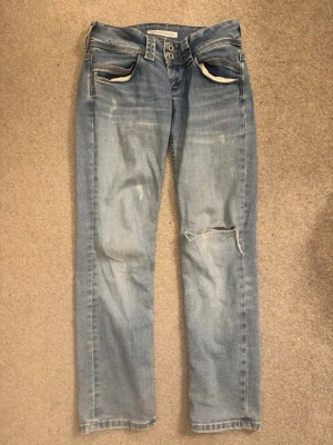 Ljusblå raka jeans från Pepe Jeans - Säljer ett par ljusblå jeans från Pepe Jeans, modell Wiser Wash. Jeansen har raka ben, slitningar och ett hål på ena knät för en trendig look. Klassisk femficksmodell med dubbla knappar i midjan. Perfekta för dig som gillar en avslappnad stil. Storlek 27/30