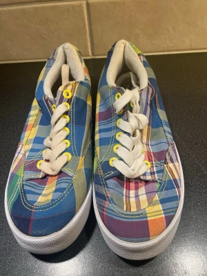 Rutiga U.S. Polo Assn.  sneakers - Färgglada US Polo Assn.  sneakers med rutigt mönster i blått, gult, rött och grönt. Skorna har vita sulor, vita snören och gula öljetter. Tillverkade i textil med klassisk låg siluett och ikonisk sidostripes. Perfekta för dig som vill sticka ut med din stil.