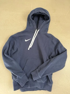 Blå Nike hoodie  - Snygg blå hoodie från Nike med vit logga på bröstet och vita dragsnören i huvan. Klassisk känguruficka framtill och ribbade muddar vid ärmar och nederkant. Perfekt för en avslappnad och sportig stil. Bra skick, pris går att diskutera!!!
