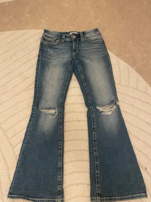 Blå bootcut jeans med slitningar - Ett par blå bootcut jeans med slitningar på båda knäna och lätt tvättad look. Jeansen har klassisk femficksdesign och låg midja. Perfekta för dig som gillar en avslappnad och trendig stil.