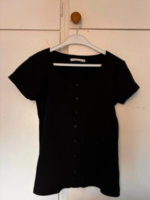 Svart ribbad topp med knappar - En svart ribbad topp med korta ärmar och knappar framtill. Toppen har en fyrkantig ringning och är i ett stretchigt material som sitter snyggt på kroppen. Perfekt att styla med jeans eller kjol. Liten i storleken. 