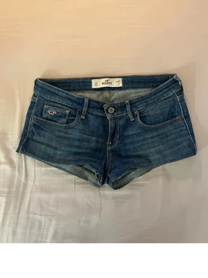 Blå jeansshorts från Hollister - Säljer ett par klassiska blå jeansshorts från Hollister med låg midja och fem fickor. Shortsen har en knappgylf och den ikoniska Hollister-loggan på fickan. Perfekta för varma dagar och enkel att matcha med allt.