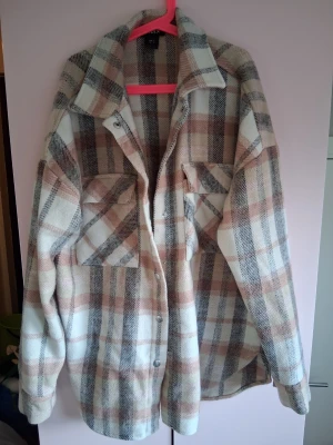 Rutig overshirt i beige och grå - Overshirt från H&M i mjukt, tjockt material med rutigt mönster i beige, vitt, grått och ljusbrunt. Två stora bröstfickor med lock och knappar framtill. Avslappnad passform och klassisk krage. Perfekt lager på lager-plagg för kyligare dagar.