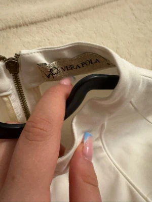 Vit klänning från Vera Pola - Snygg vit klänning från Vera Pola med hög krage och dragkedja i ryggen. Klänningen har en stilren look och är tillverkad i ett slätt tyg som ger en elegant känsla.