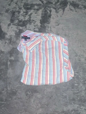 Tommy Hilfiger Striped shirt  - Sjukt fräsch randig skjorta från Tommy Hilfiger i en unik färgkombination med pastellfärger: blått, rosa och vitt (se närbild sista) den perfekta skjortan för våren och sommaren! • Passform: Klassiskt snitt. Funkar lika bra att bära som den är eller öppen över ett linne för en avslappnad look. • Material: Lätt och skönt tyg (seersucker-liknande struktur på närbilden) som andas bra och håller dig sval. • Skick: Toppskick, nytvättad och redo att
