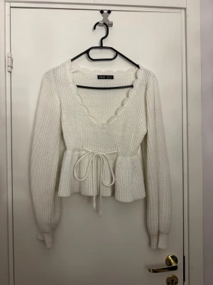 Vit stickad topp från Shein - Supersöt vit stickad topp från Shein med volangkantad V-ringning och knytband i midjan. Tröjan har lång ärm, ribbstickade muddar och en peplumliknande form nedtill. Perfekt för dig som vill ha en feminin och trendig look.