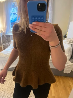 Brun stickad topp med volang ifrån h&m - Säljer en brun stickad topp med korta ärmar och volang nedtill. Tröjan har en mjuk, fluffig struktur och sitter figurnära upptill. Har används kanske 2-3 ggr.