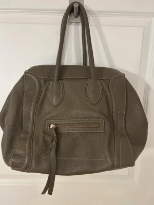 Grågrön handväska i skinn - Snygg grågrön handväska i mjukt skinn med dubbla handtag och dekorativ dragkedja framtill. Väskan har en rymlig form och sväljer mycket samtidigt som den är smidig att bära. Mkt fint skick!