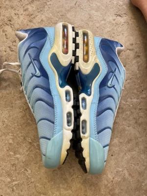 Nike Air Max Plus blå sneakers - Nike Air Max Plus sneakers i olika blå nyanser med mesh och syntetmaterial. Skorna har synliga Air Max-bubblor i sulan, svarta och vita detaljer samt den ikoniska Swoosh-loggan på sidan. Sulan har ett unikt mönster och Tuned Air-detaljer. Perfekta för dig som gillar streetwear.