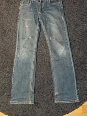 Blå raka jeans från Levi's - Säljer ett par klassiska blå jeans från Levi's med raka ben och slitna detaljer på framsidan. Jeansen har fem fickor, normal midja och en snygg tvättad look. Perfekta för en avslappnad stil och passar till det mesta i garderoben.