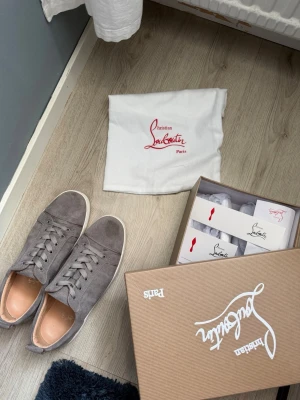 Gråa sneakers från Christian Louboutin - Snygga gråa sneakers från Christian Louboutin i mocka med vita sulor och klassisk låg profil. Skorna har matchande grå skosnören och diskreta detaljer som ger en clean look. Perfekta för dig som gillar exklusiva och stilrena sneakers.