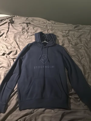 Mörkblå DIF hoodie Stockholm - Snygg mörkblå hoodie från Djurgårdens IF med klubbens logga och texten STOCKHOLM på bröstet. Klassisk känguruficka framtill och justerbar huva med dragsko. Perfekt för dig som vill visa ditt stöd för DIF och hålla stilen.