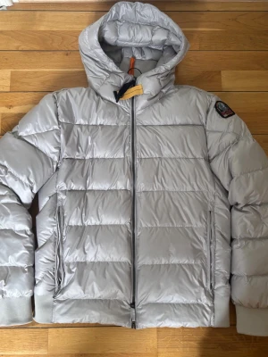 Silver pufferjacka från Parajumpers - Snygg silverfärgad pufferjacka från Parajumpers med huva och dragkedja framtill. Jackan har två sidofickor med dragkedja, ribbade muddar och en patch med logga på ärmen. Perfekt för kalla dagar och riktigt trendig look.