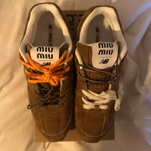 Bruna Miu Miu x New Balance sneakers - Unika bruna sneakers i mocka från Miu Miu x New Balance med vit, orange och brun snörning. Skorna har vit vadderad insida och tydlig branding på plösen. Snygg retrostil och cool mix av färger på skosnörena gör dem till ett statement i garderoben.