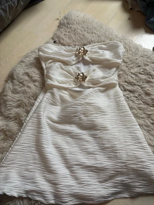 Vit axelbandslös kortklänning med blommor - Säljer en vit axelbandslös kortklänning med snygg struktur i tyget och två guldfärgade blomdetaljer framtill. Klänningen har en omlottliknande twist vid bysten och är perfekt för sommarens alla tillfällen.