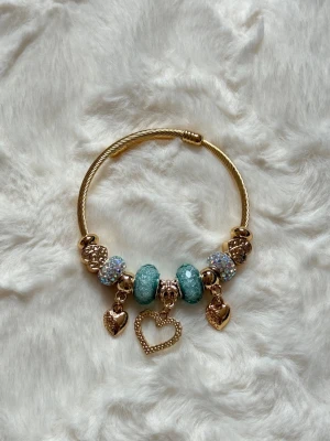 Guldarmband med pärlor och hjärtan - Snyggt armband i guldfärgad metall med glittriga pärlor i turkost och silver. Armbandet har flera berlocker, bland annat tre hjärtan och dekorativa detaljer. Perfekt accessoar för att lyfta din outfit.