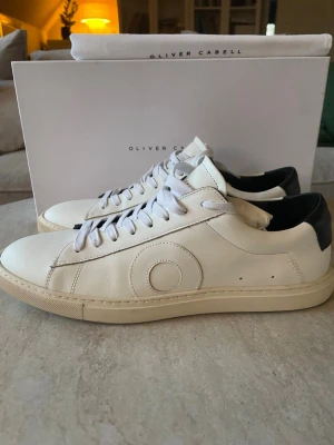 Vita Oliver Cabell sneakers i läder - Stilrena vita sneakers från Oliver Cabell i mjukt läder med svart detalj på hälen. Klassisk låg modell med vita skosnören och diskret cirkeldetalj på sidan. Perfekta för dig som gillar minimalistisk och clean stil. Storlek 42 men passar 43🙌